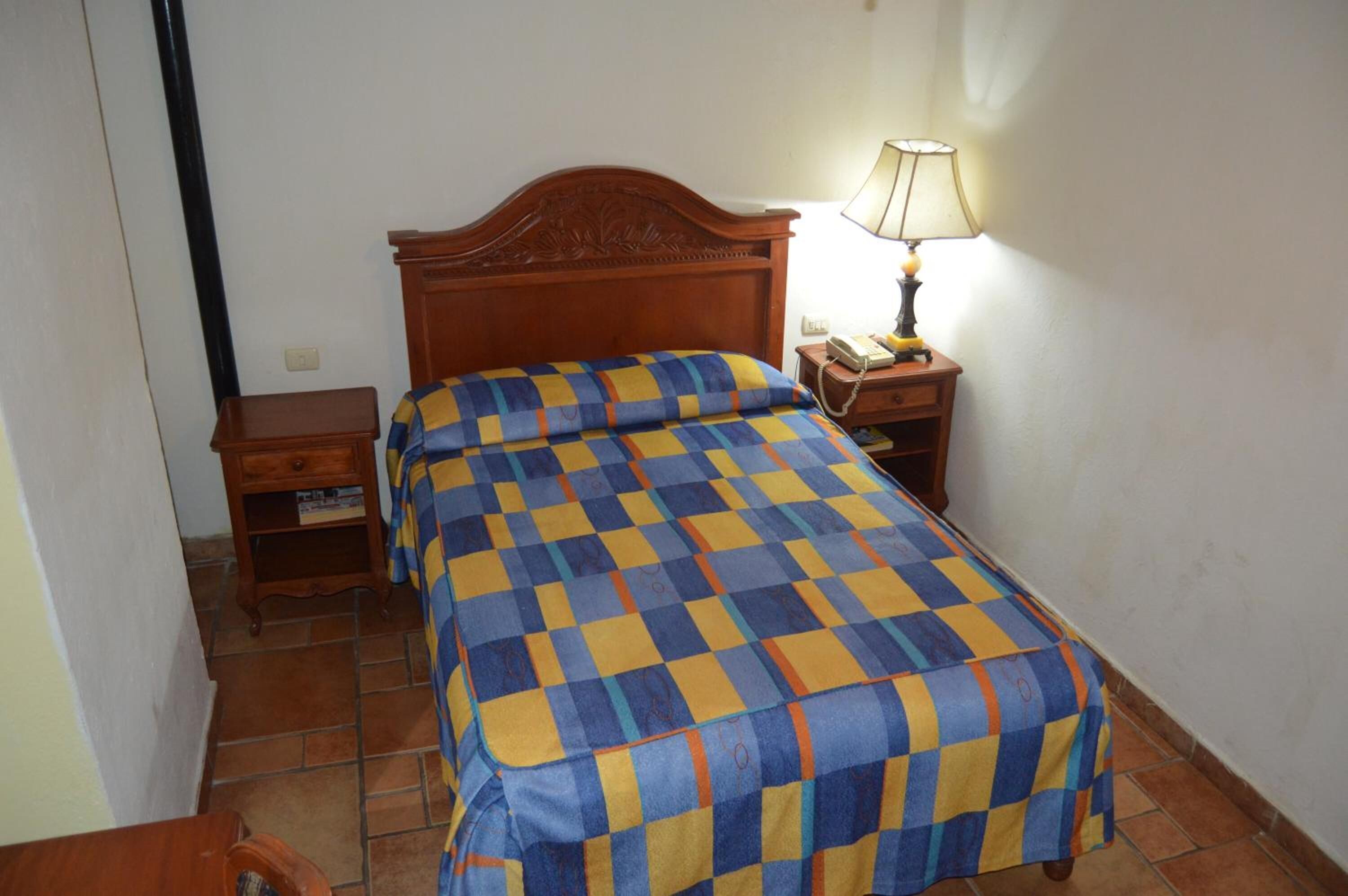 A single bed with a checkered bedspread, a wooden headboard, a bedside table with a lamp, and a small wooden nightstand.