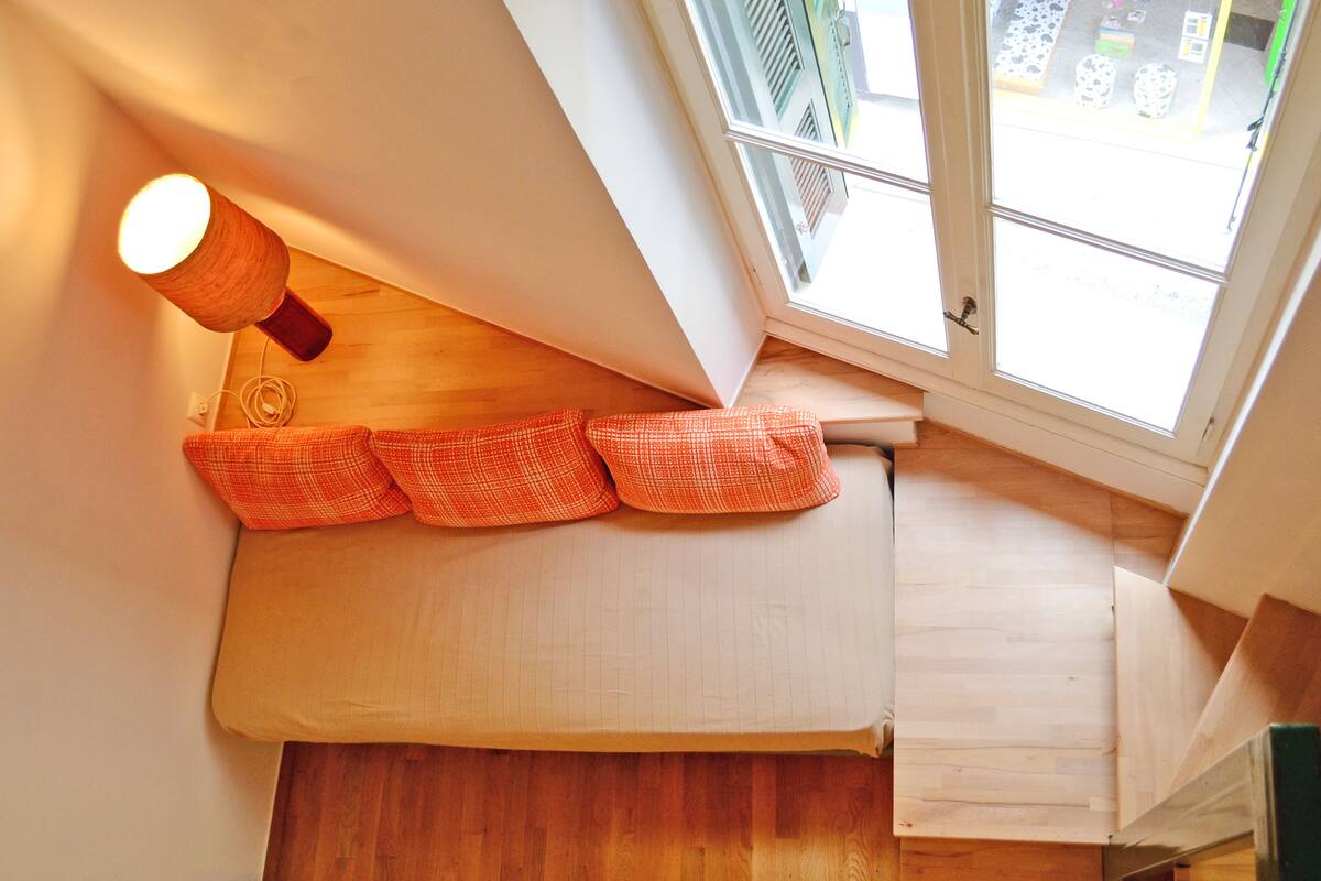 Une pièce aux hauts plafonds, dont un est en pente, avec un plancher en bois, une fenêtre donnant sur la rue et une petite table sur laquelle se trouve une lampe.