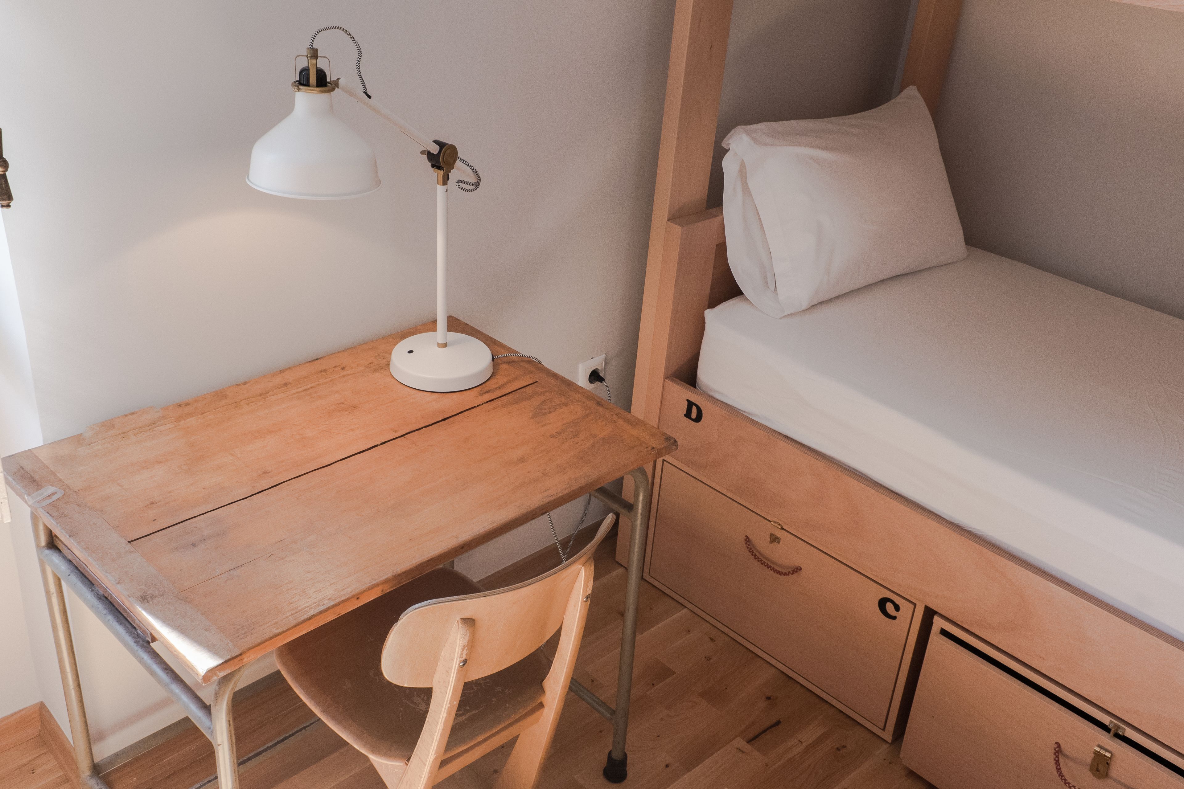A small, compact room with a wooden desk, a chair, a single bed with a white mattress and pillow, and a white desk lamp.