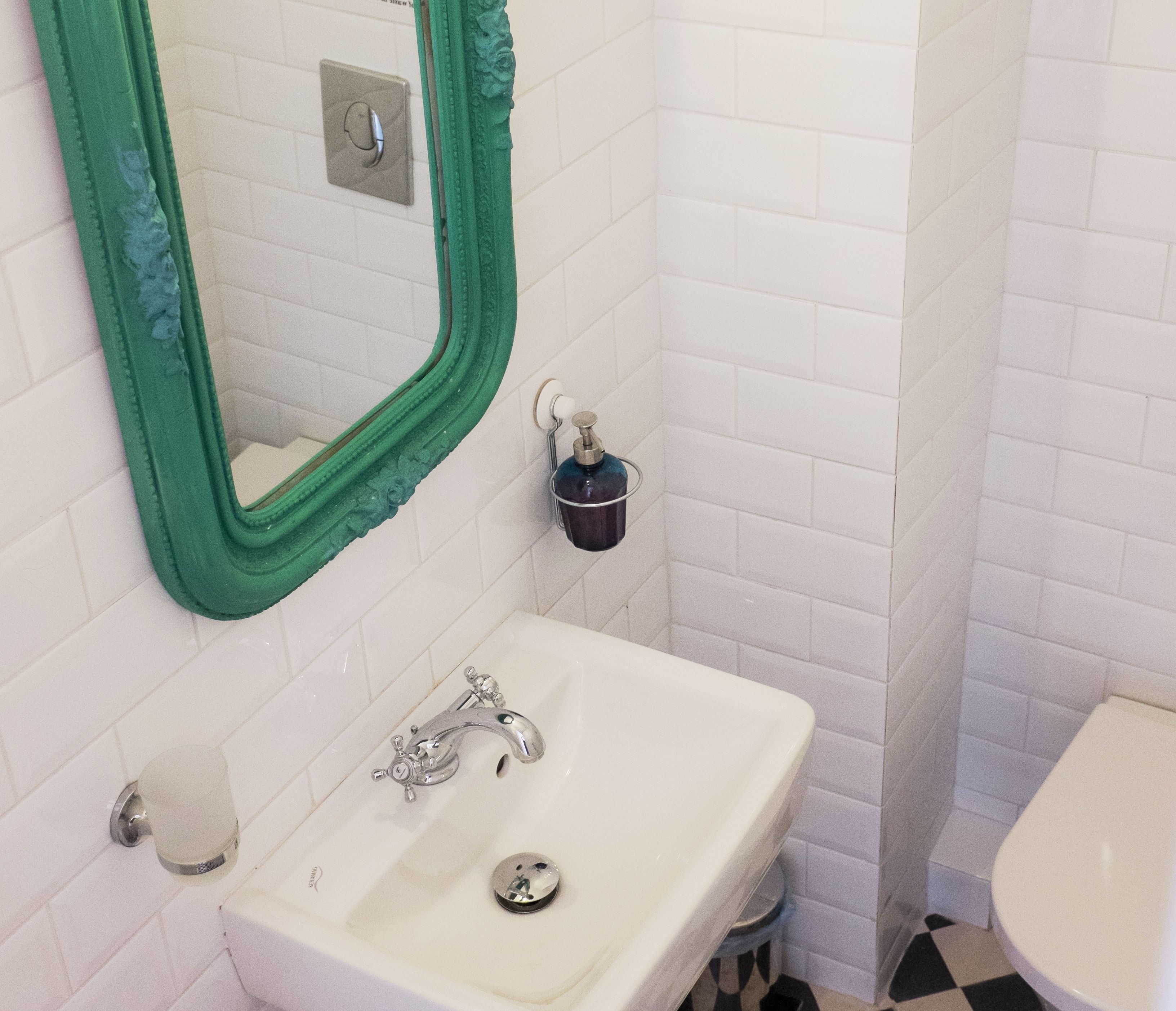 A bathroom with a white sink, a green-framed mirror, a small bucket, and a toilet.