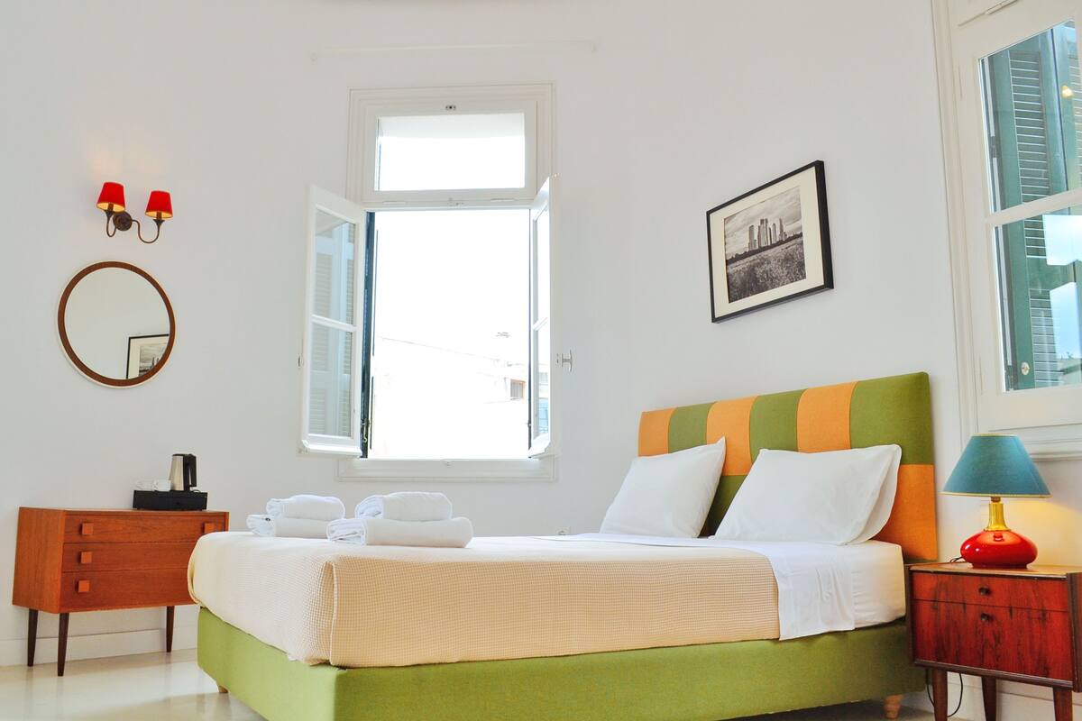 Une chambre à coucher bien rangée, avec un lit, une commode, un miroir et une fenêtre dont le volet est ouvert.
