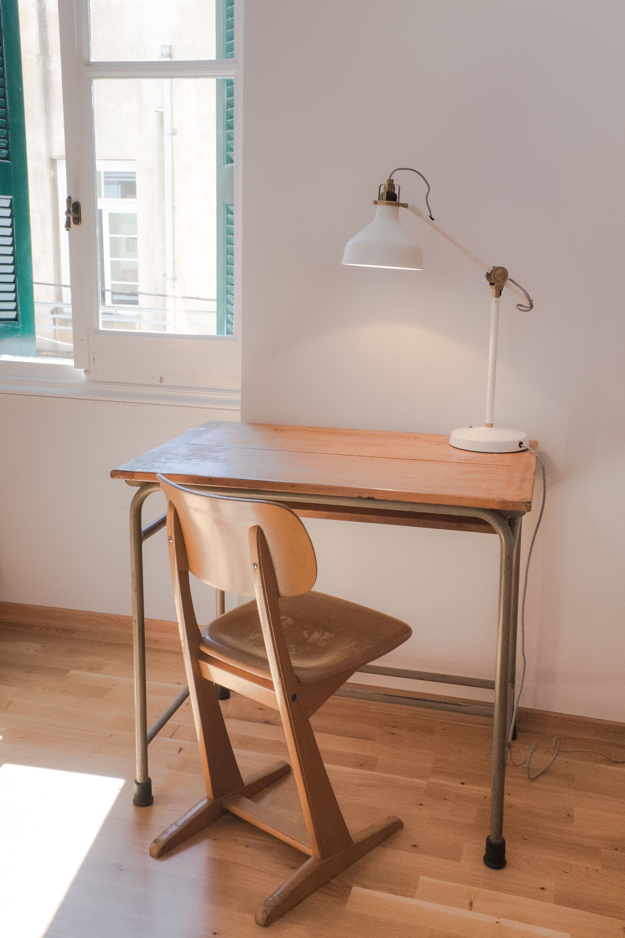 Een bureau met een stoel, een witte bureaulamp en een raam met jaloezieën.