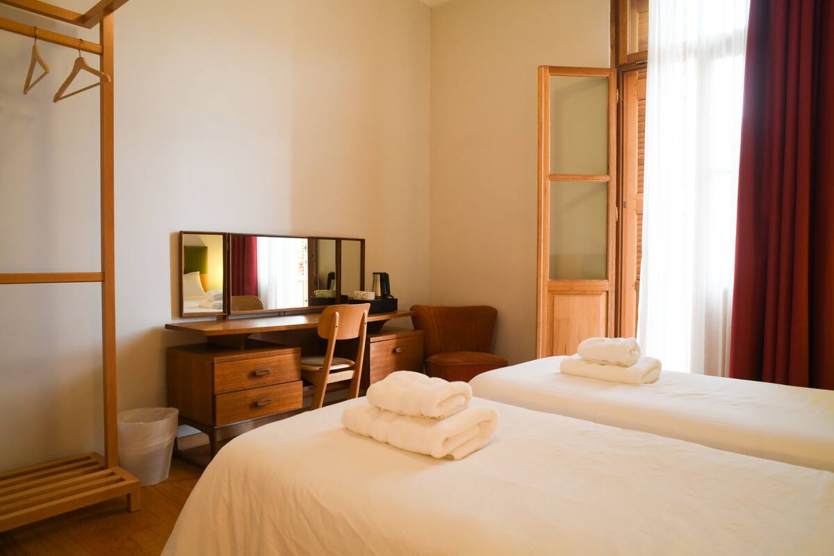 Une chambre d’hôtel avec deux lits, un bureau en bois, une chaise, un miroir et une fenêtre avec des rideaux.