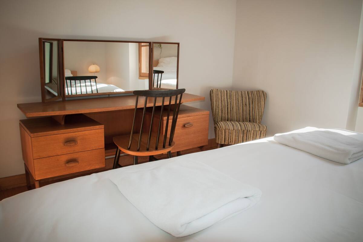 Une chambre d’hôtel avec un lit, un bureau en bois doté de tiroirs, une chaise, un fauteuil à rayures et un grand miroir.