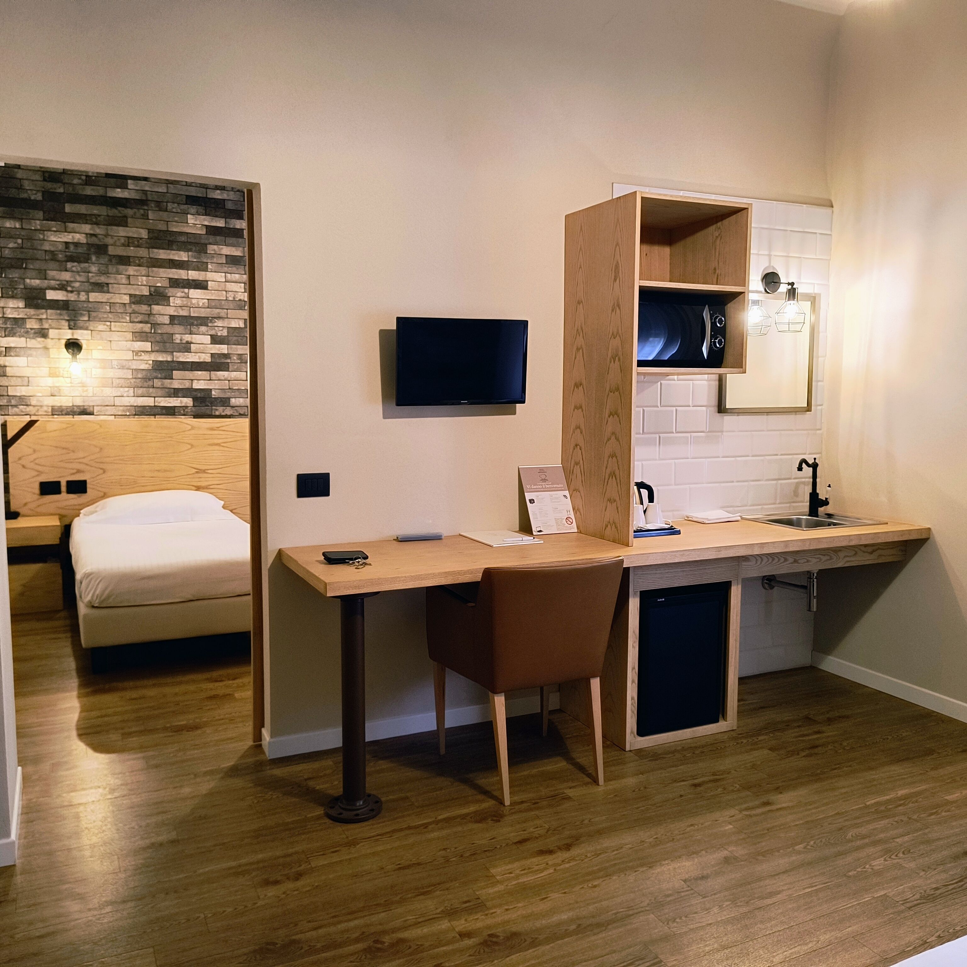 A compact hotel room with a wooden desk, a chair, a bed, a TV, a small kitchenette, and a brick-patterned wall.
