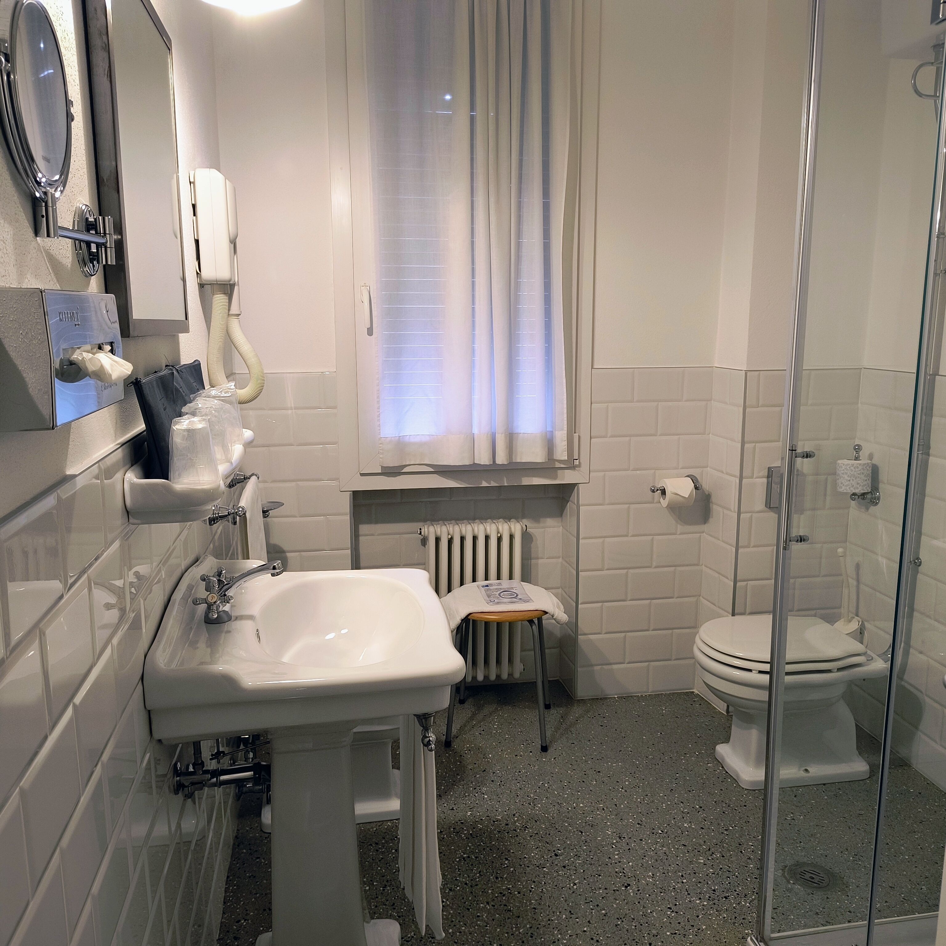 A modern bathroom with a white sink, toilet, and shower. There is a towel rack, a mirror, and a window with curtains.