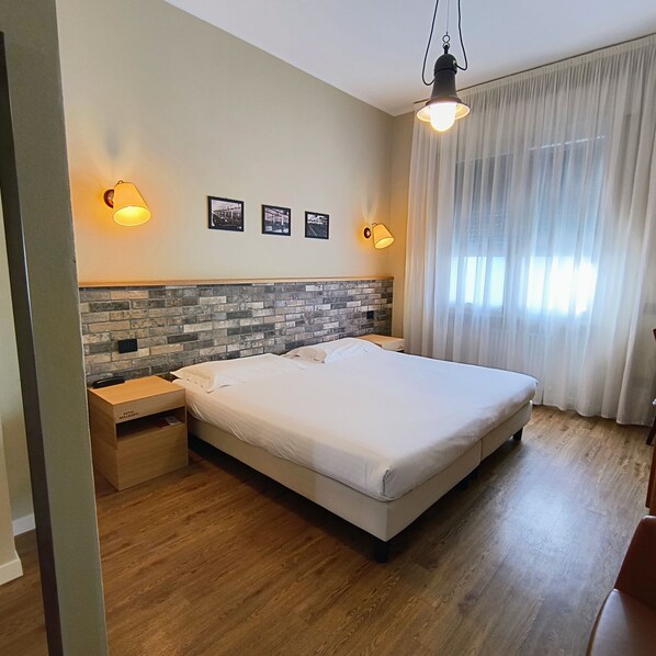 A neatly arranged hotel room with a large bed, two bedside tables, a brick-patterned headboard, and a window with sheer curtains.