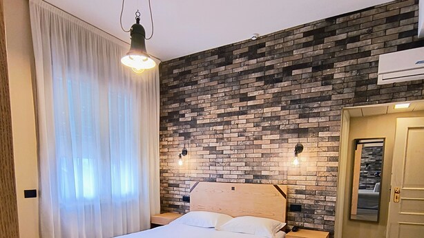 A modern bedroom with a wooden bed, white curtains, a brick-patterned wall, and a ceiling light. - Hotel Miramonti (Schio)