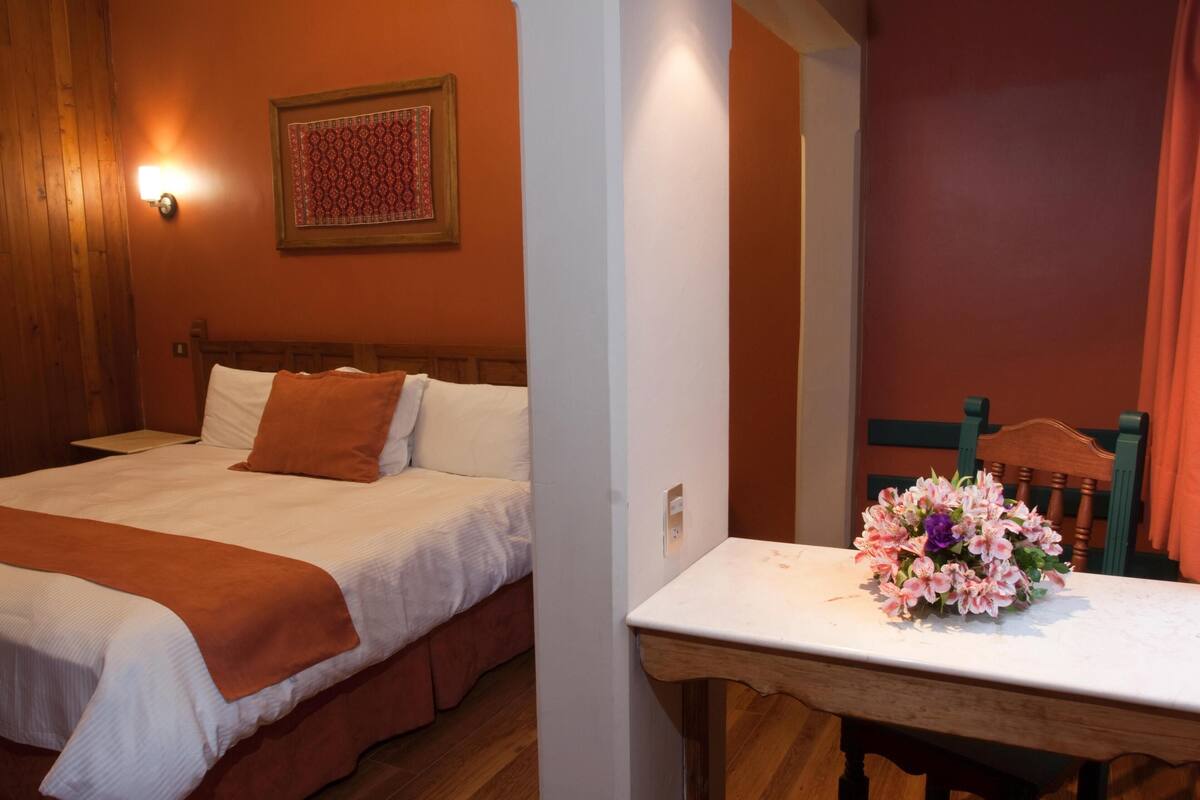 Une chambre à coucher comprenant un lit, une table de chevet, une tête de lit en bois, une lampe fixée au mur et une tapisserie encadrée.