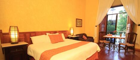 A hotel room with a large bed, two bedside tables with lamps, a wooden headboard, a small table with a chair, and a window with curtains.