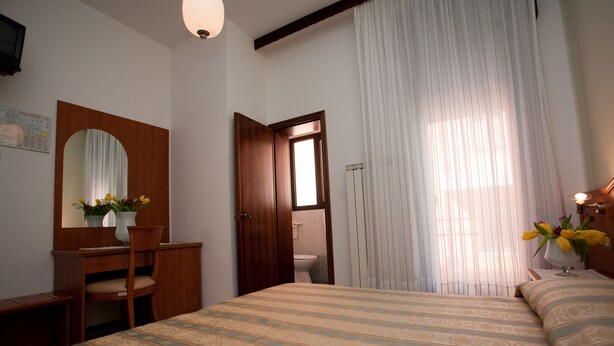 A bedroom with a bed, a wooden dresser, a chair, a mirror, and a window with curtains. - Hotel Ideal (Chioggia)