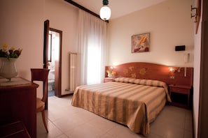 A hotel room with a bed, a wooden nightstand, a chair, a desk with a lamp, a vase with flowers, a painting on the wall, and a door leading to a bathroom.