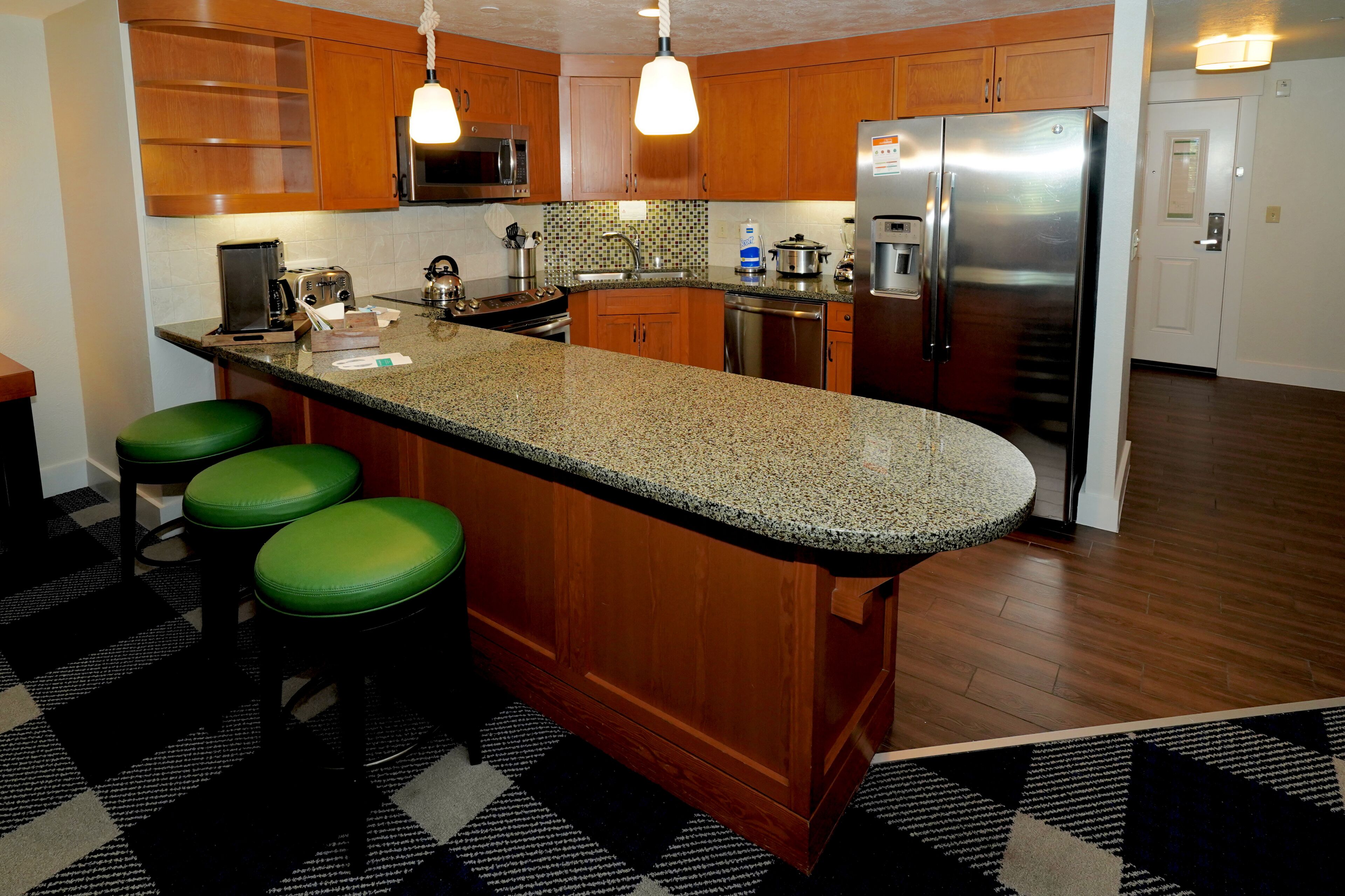 Una cocina con armarios de madera, encimera de granito, refrigerador de acero inoxidable y taburetes de bar verdes.