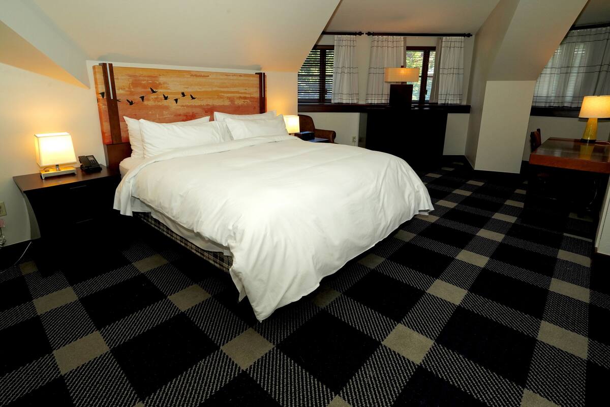 A hotel room with a large bed, a wooden headboard, a checkered floor, a desk with a lamp, and a window with curtains.