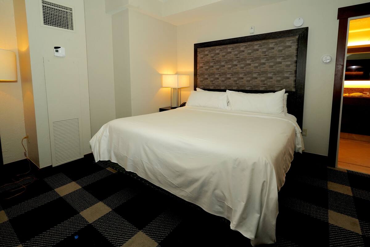 A hotel room with a large bed, a nightstand with a lamp, a wall-mounted air conditioning unit, and a checkered floor.