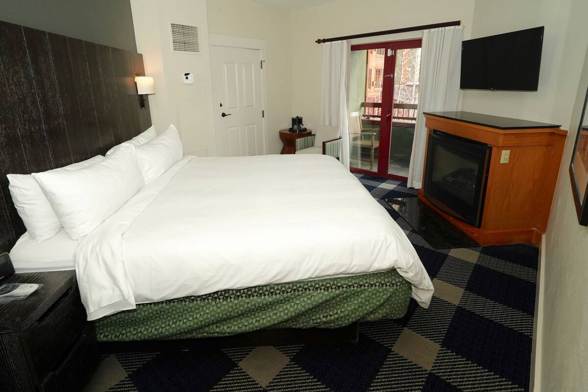 A hotel room with a large bed, a television, a small table with a lamp, and a sliding glass door to a balcony.
