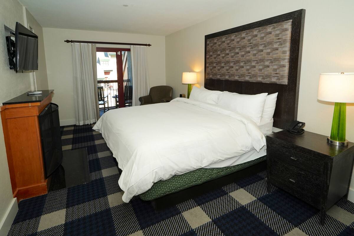 A hotel room with a large bed, a television mounted on the wall, a desk with a telephone, and a chair.