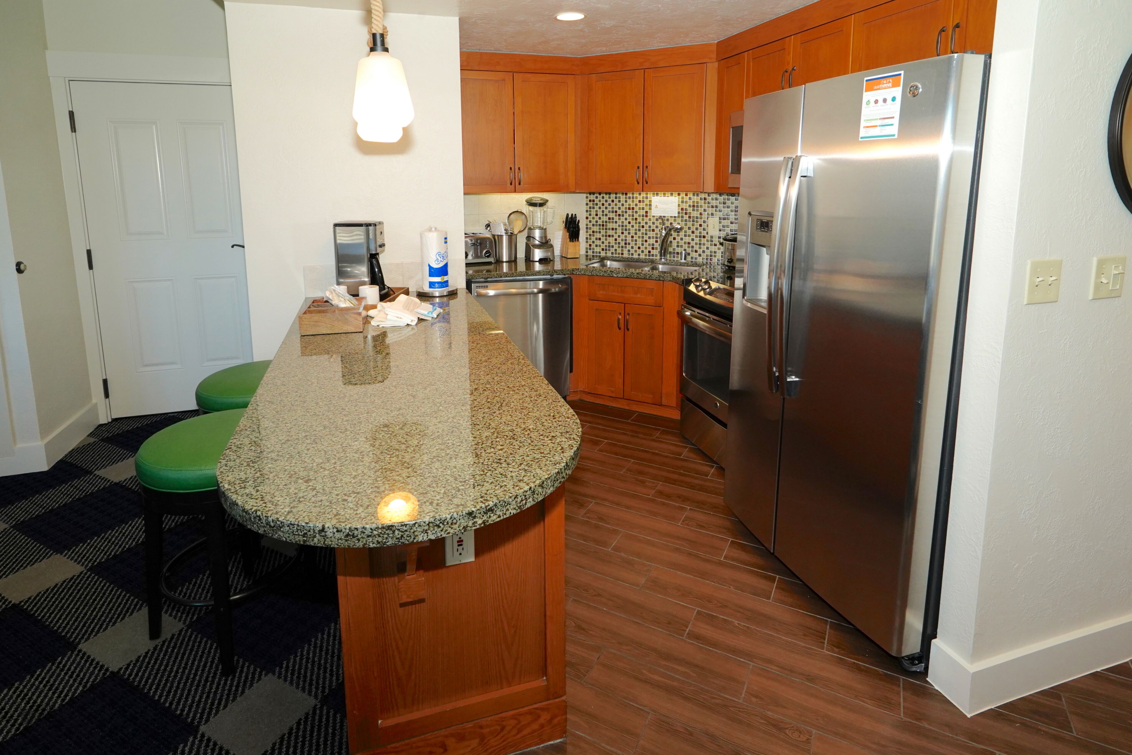 A kitchen with wooden cabinets, a stainless steel refrigerator, a granite countertop, and a green stool.