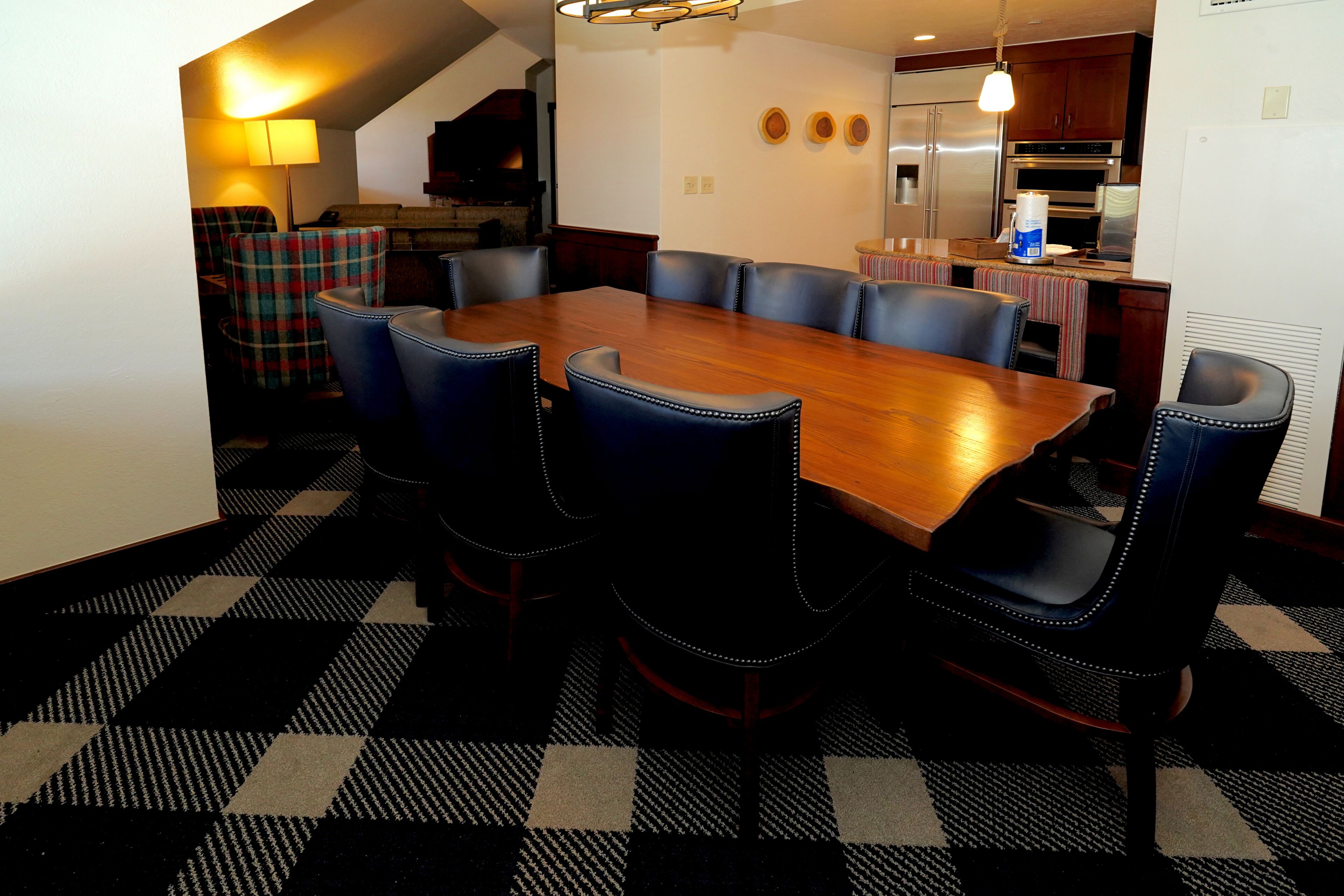A dining area with a large wooden table surrounded by six chairs, a checkered floor, and a kitchen in the background.