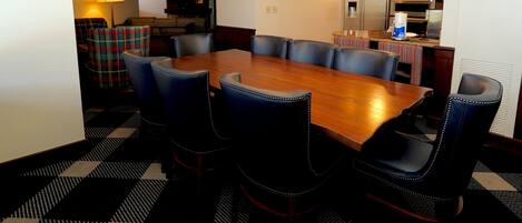 A dining area with a large wooden table surrounded by six chairs, a checkered floor, and a kitchen in the background.