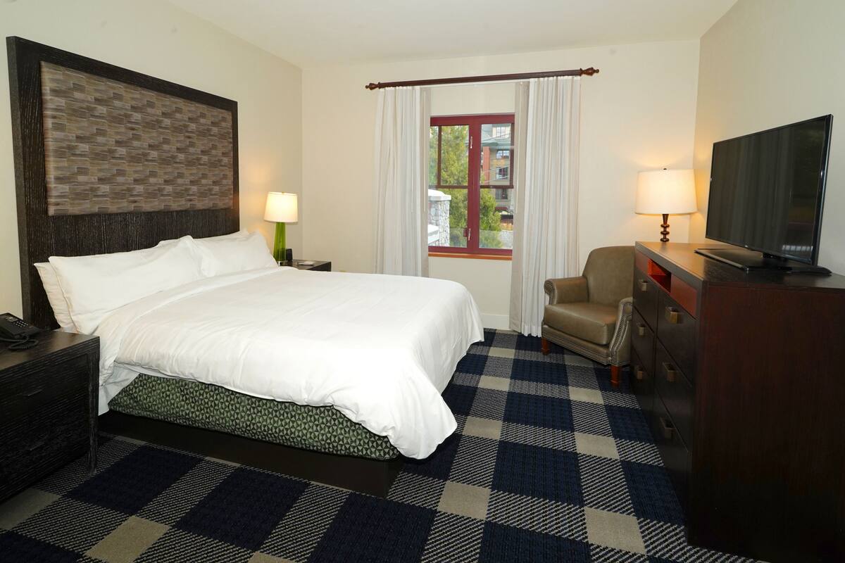 A hotel room with a large bed, a television, a desk with drawers, a chair, and a window with curtains.
