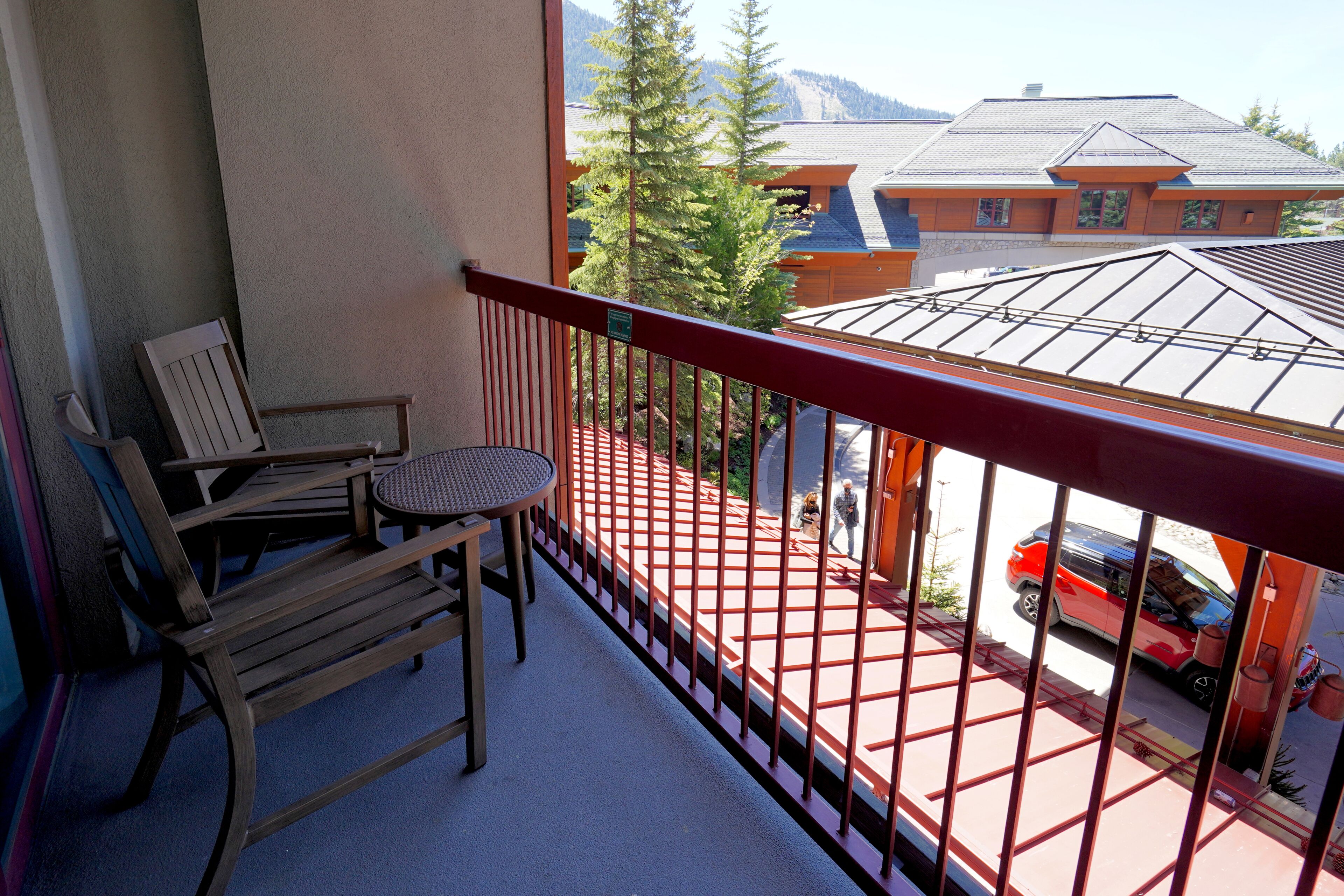 A balcony with wooden chairs and a small table, overlooking a street with a red car and buildings in the background.