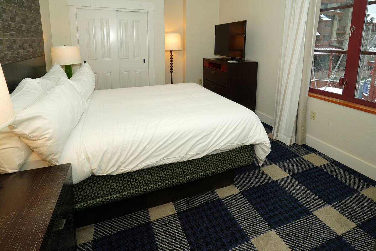 A hotel room with a bed, a desk, a TV, a lamp, and a patterned rug.