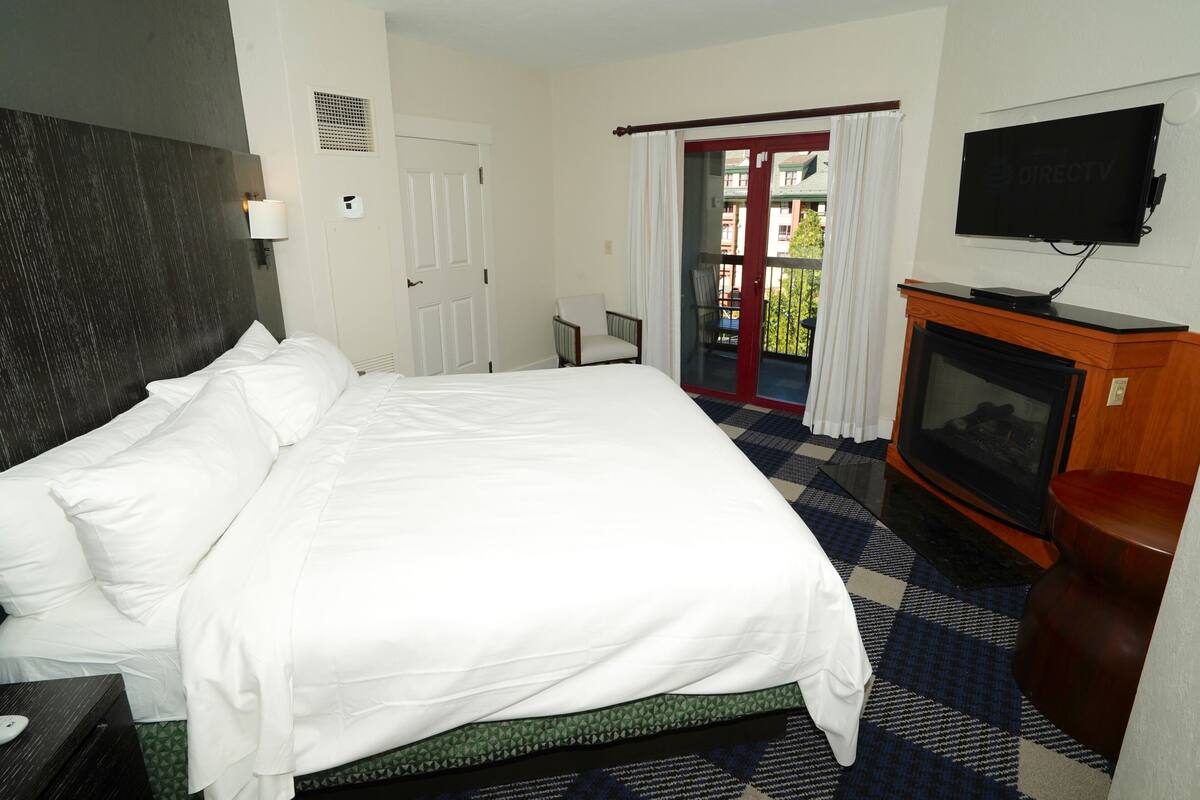 A hotel room with a large bed, a TV, a fireplace, and a balcony with a view.