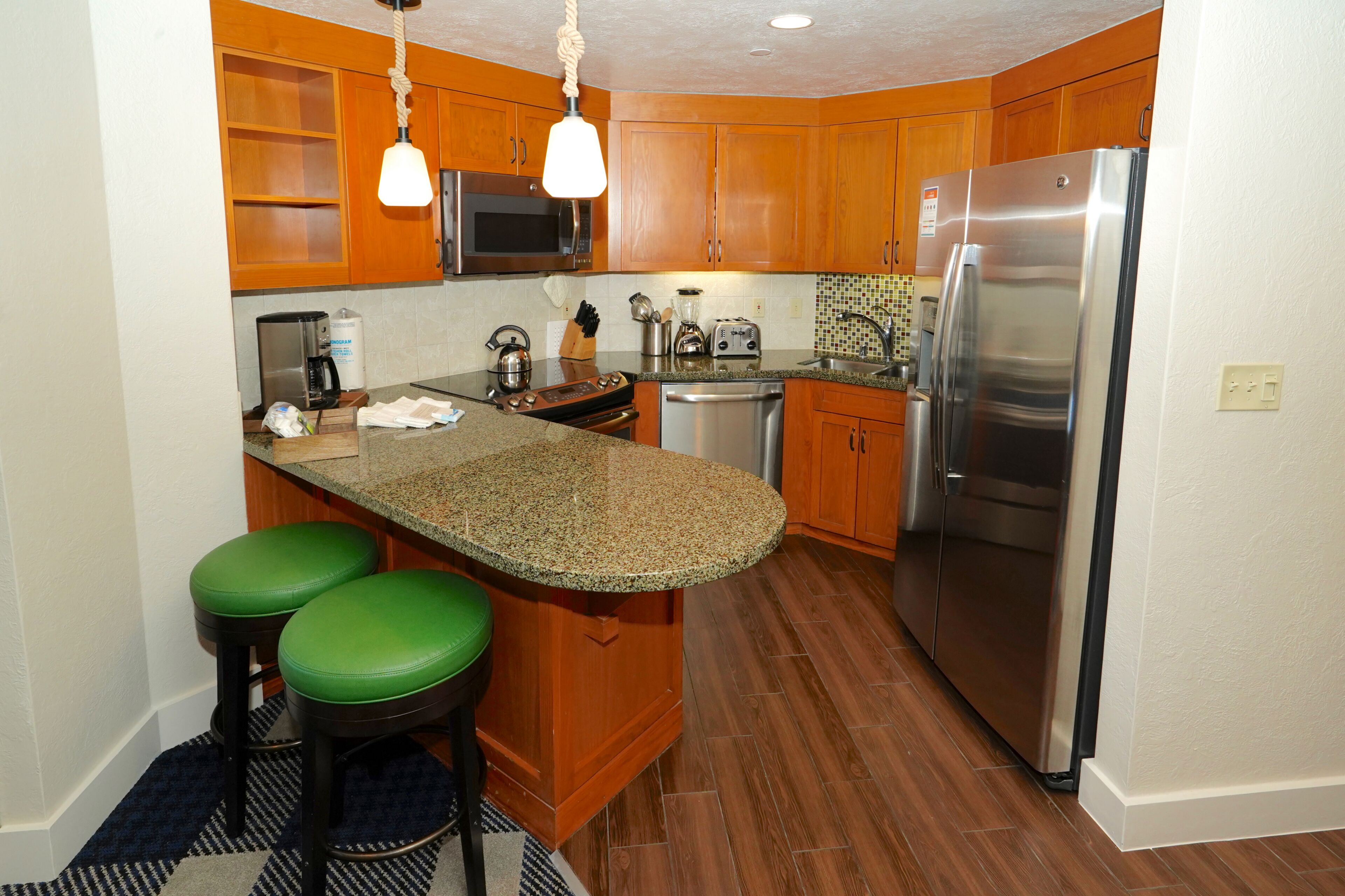 A kitchen with wooden cabinets, a stainless steel refrigerator, a microwave, a stove, and two green bar stools.