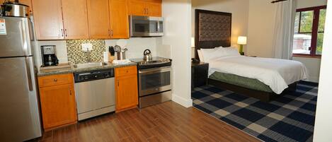 A compact hotel room with a kitchenette, a bed with a checkered area rug, and a window with curtains.
