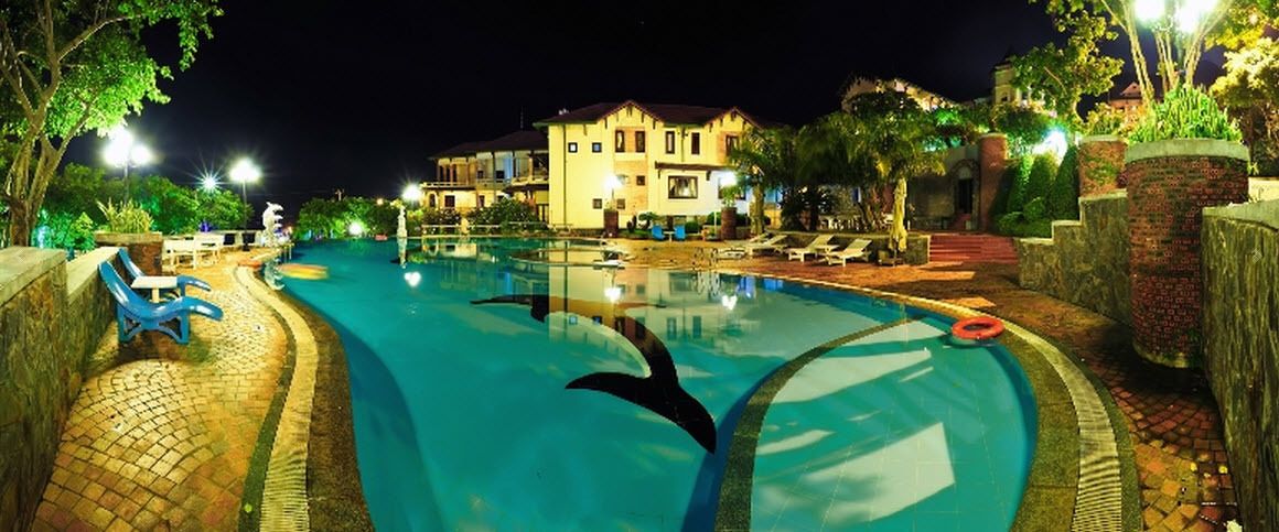 Un espace piscine éclairé la nuit, avec des chaises longues, un bâtiment en arrière-plan et un chemin pavé de briques.
