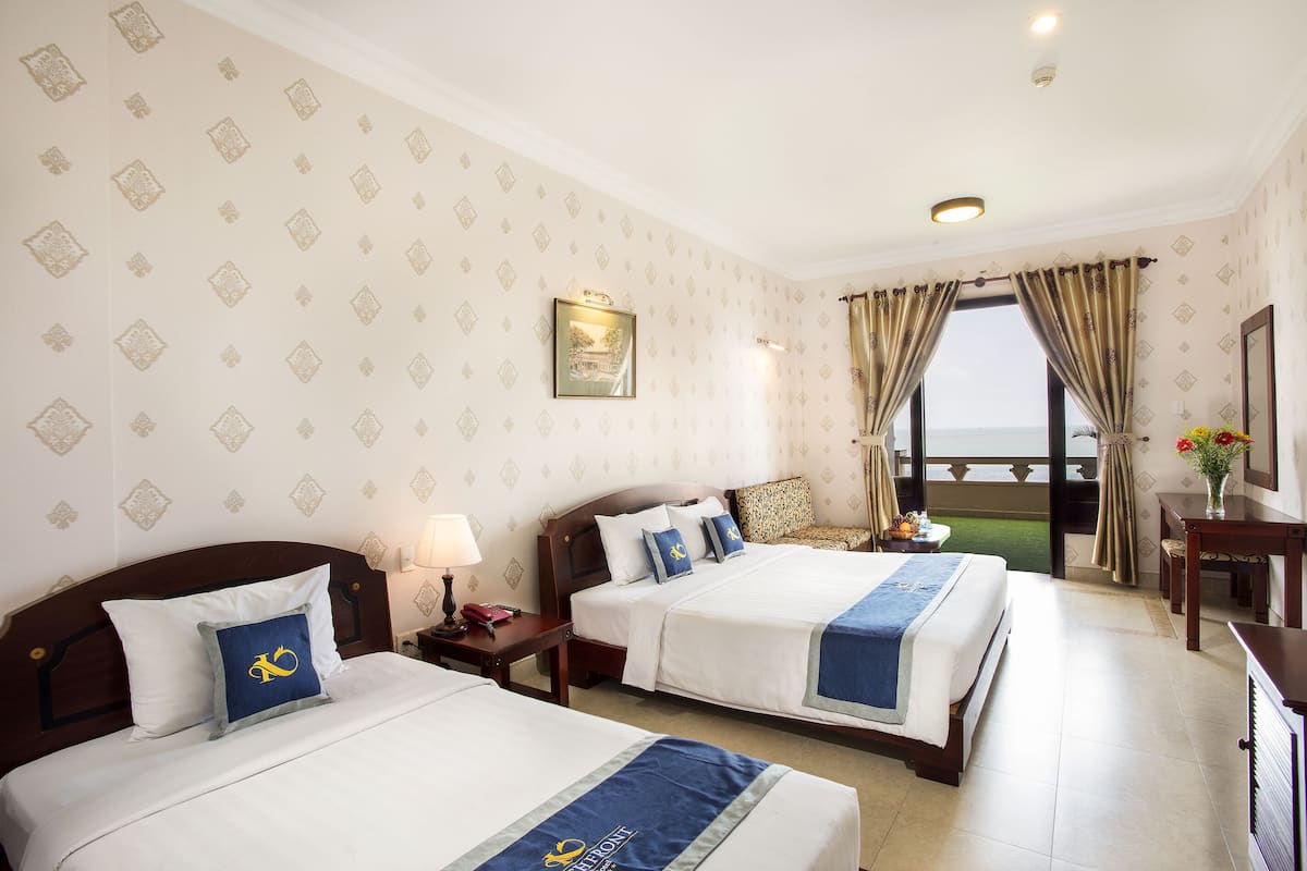 A hotel room with two beds, a nightstand, a lamp, a framed picture on the wall, and a view of the sea through the window.