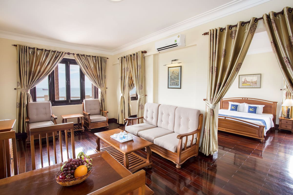 A hotel room with a sofa, chairs, a dining table with fruit, a bed, and curtains.
