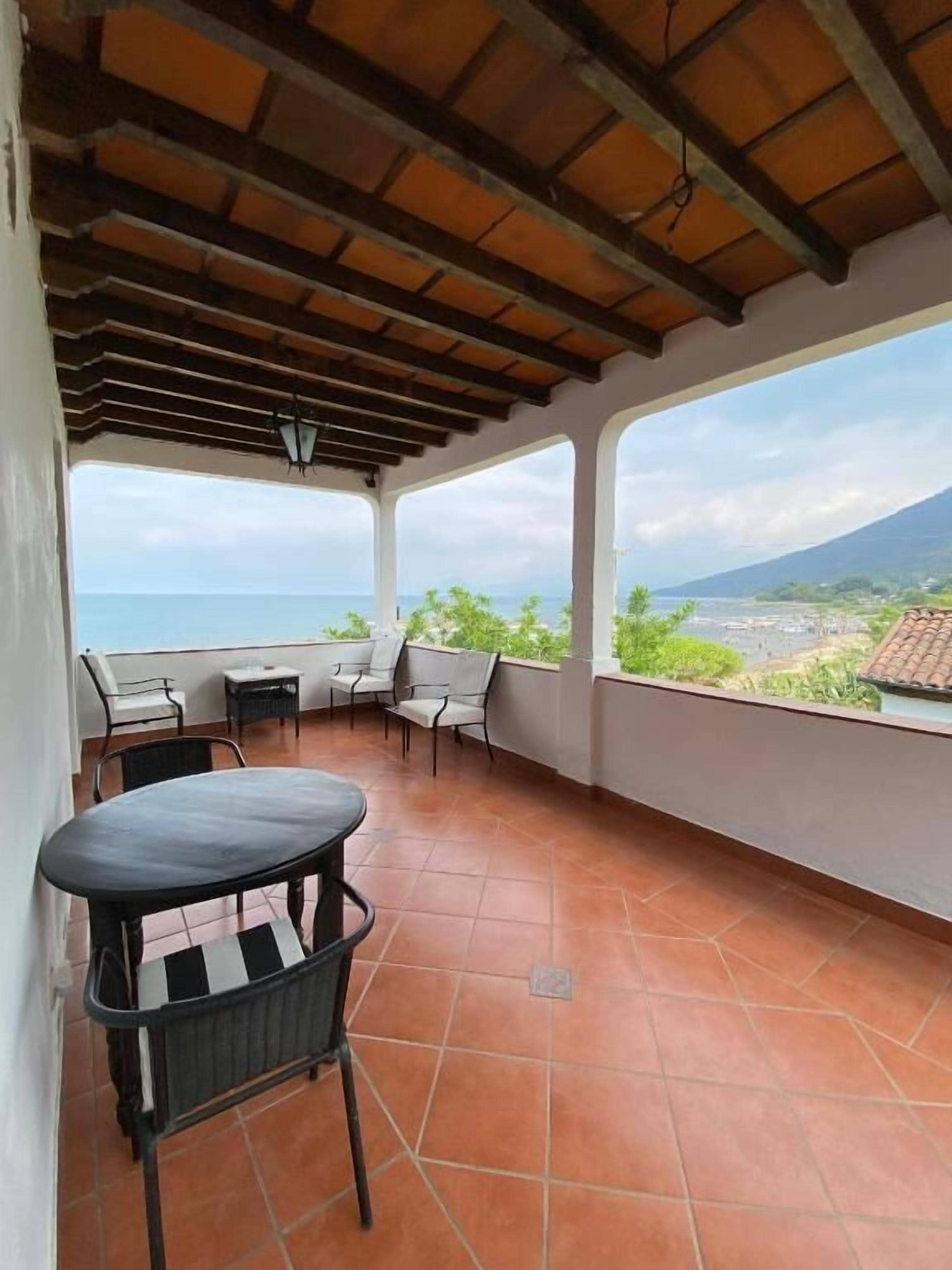 A covered outdoor area with a table and chairs, a view of the sea, and a mountain in the distance.