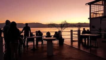 A sunset view from a terrace with tables and chairs, overlooking a body of water and distant mountains.