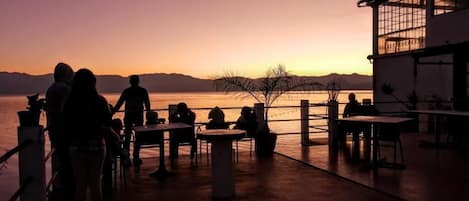 Vista al atardecer desde una terraza con mesas y sillas, con vista a un cuerpo de agua y montañas distantes.