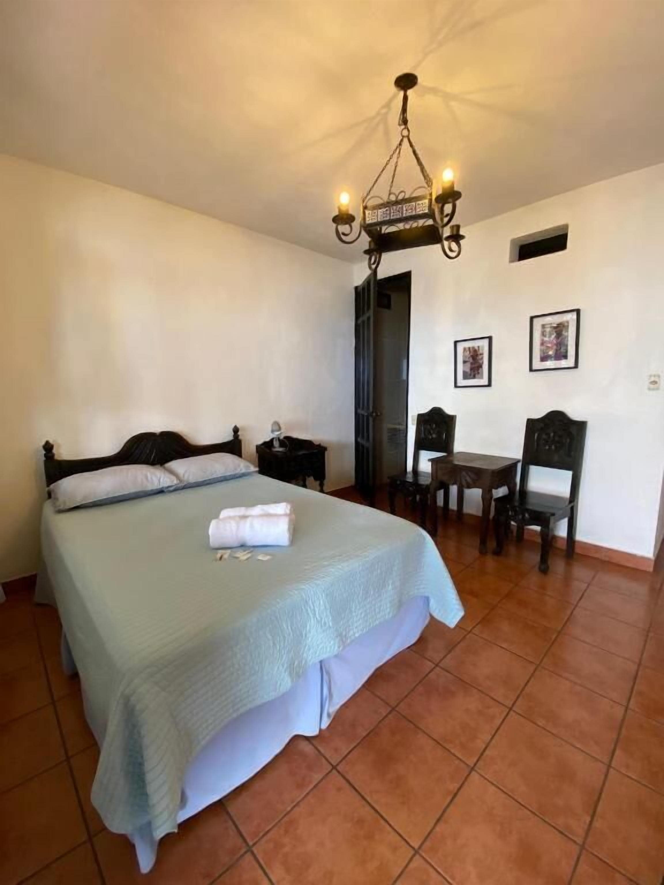 Une chambre à coucher comprenant un lit, un bureau, deux chaises, une petite table et un luminaire fixé au plafond.