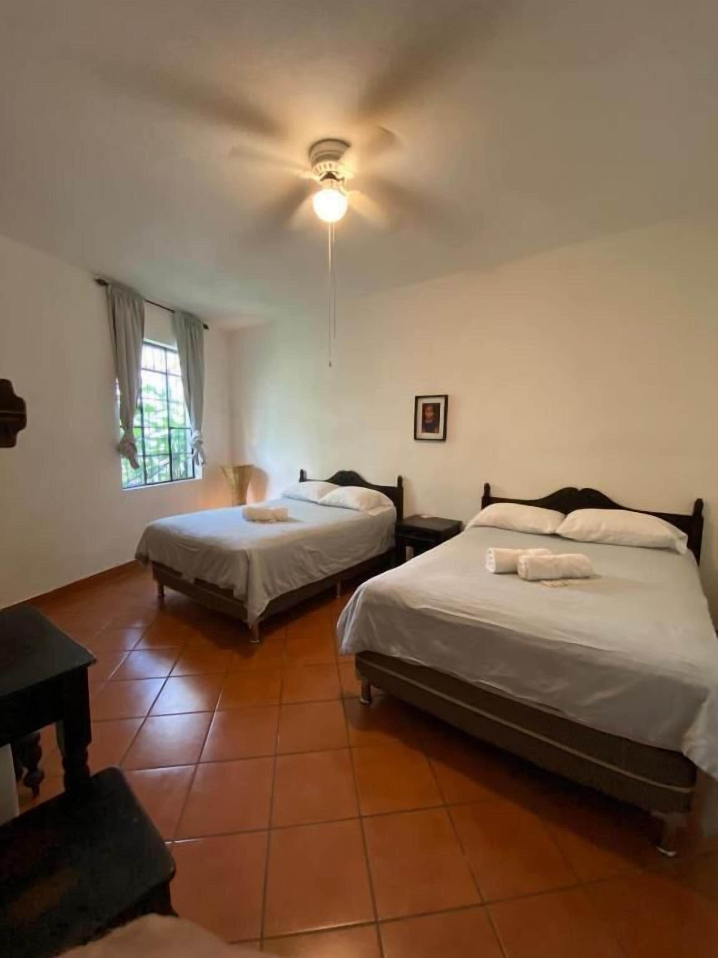 Una habitación con dos camas, un ventilador de techo, una ventana con cortinas y una pequeña mesa.