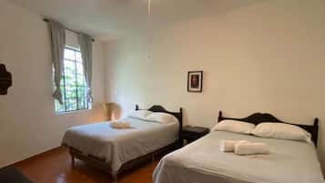 Una habitación con dos camas, un ventilador de techo, una ventana con cortinas y una pequeña mesa.