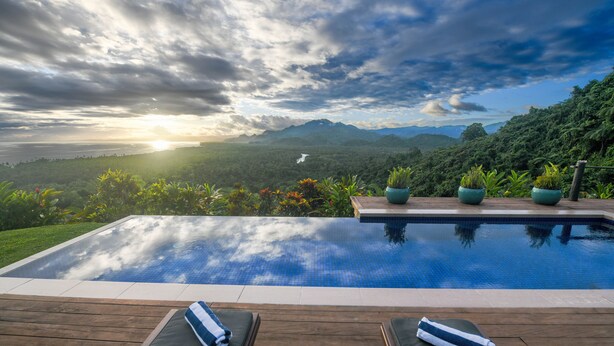 A swimming pool with wooden deck and lounge chairs, surrounded by lush greenery and mountains. - Emaho Sekawa Resort (Savusavu)