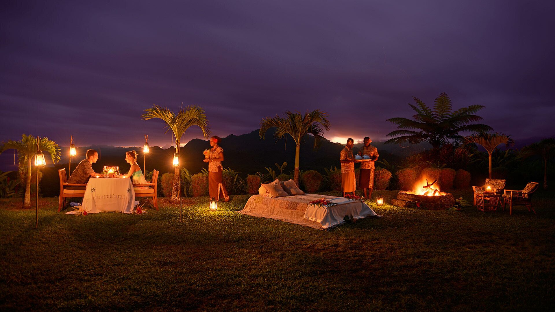 An outdoor dining area with tables and chairs, surrounded by palm trees and a fire pit.
