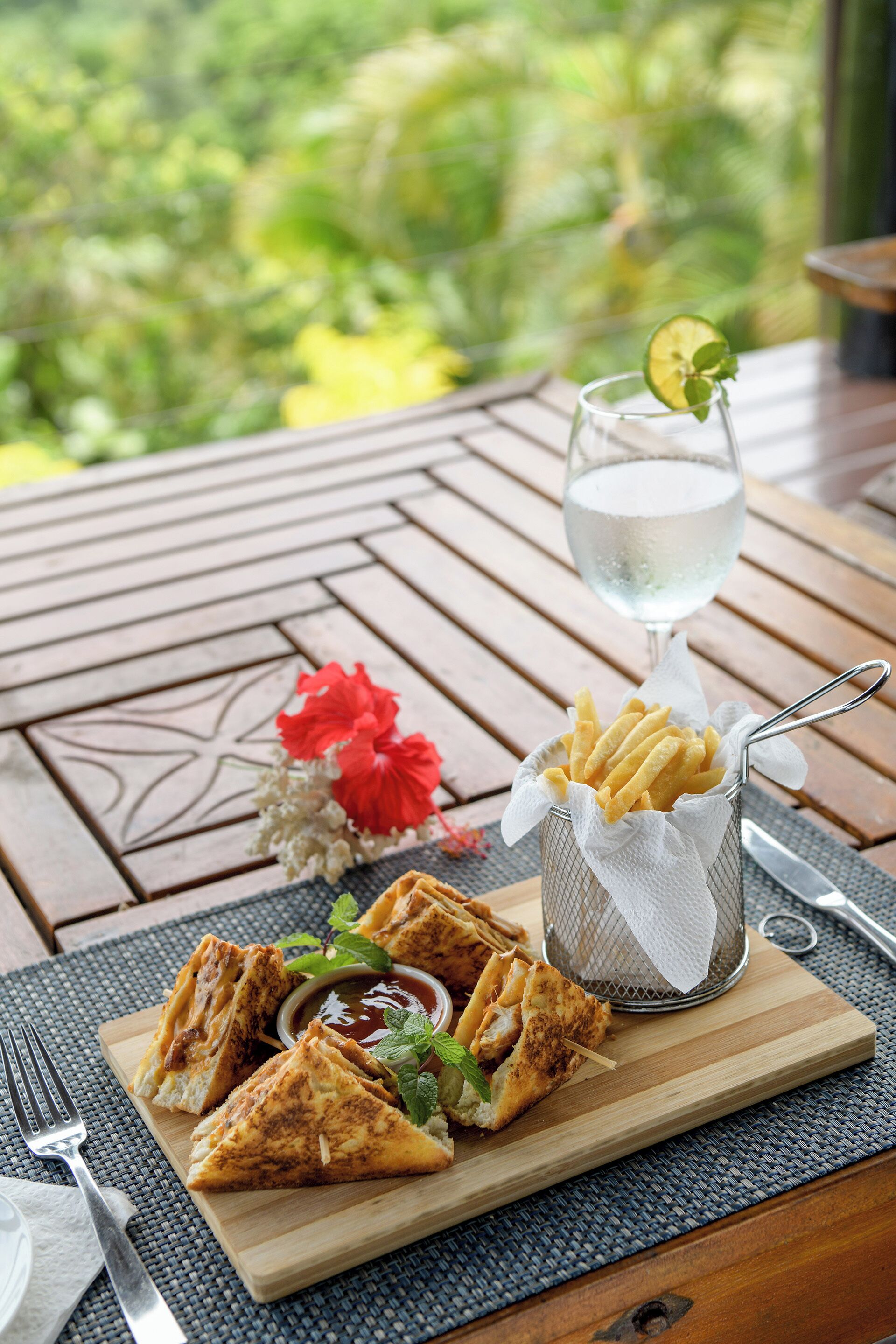 Una mesa de madera con una comida que incluye sándwiches de queso a la plancha, papas fritas y un vaso de agua, ubicada en una terraza con vistas al verdor.