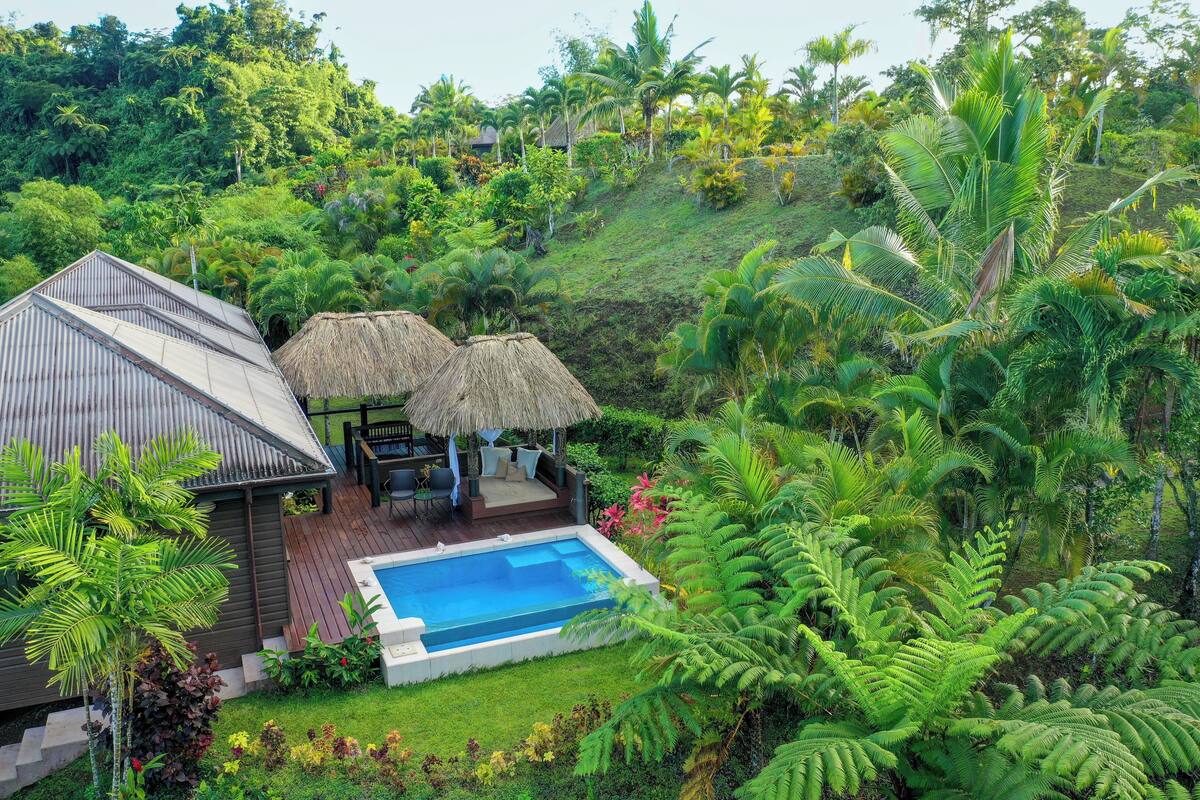 Uma casa com piscina, cercada por vegetação exuberante e uma construção com telhado de palha.