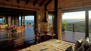 A spacious room with a wooden deck, a dining area, and a large window offering a view of the ocean and hills. - Emaho Sekawa Resort (Savusavu)