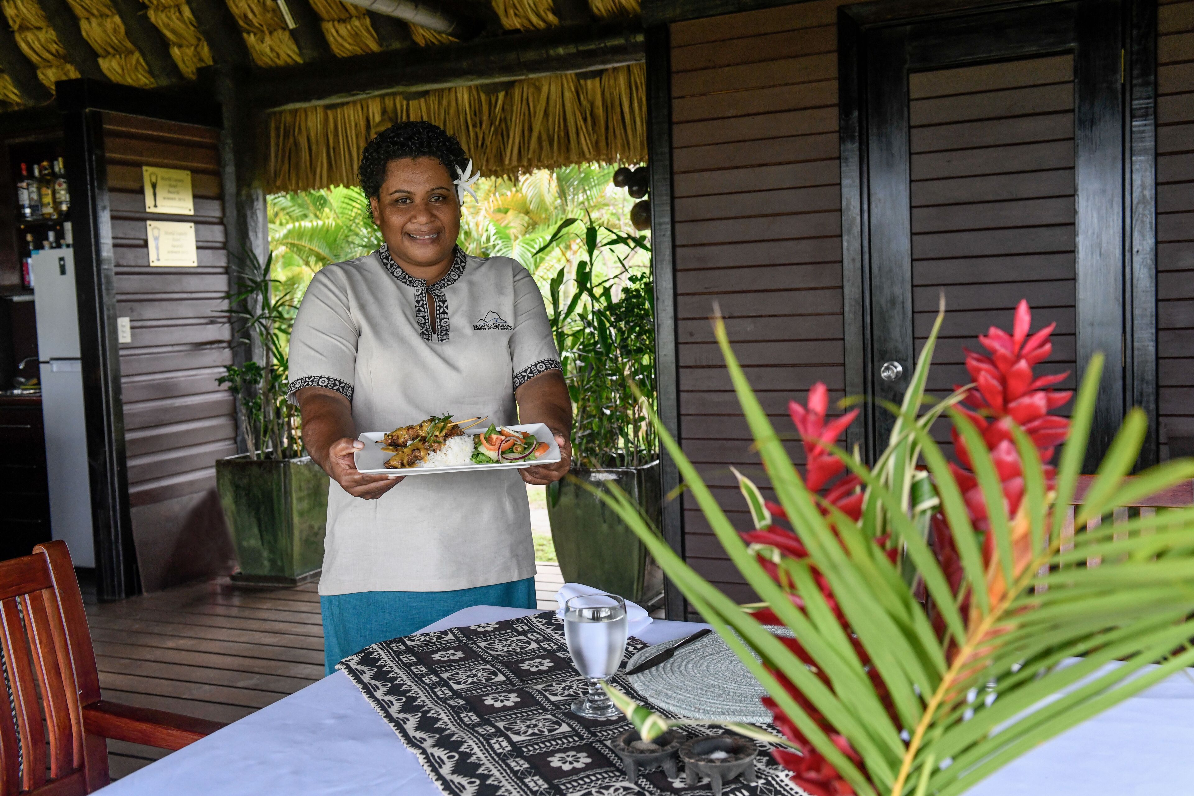 A person holding a plate of food at a table with a patterned tablecloth, a glass, and a small bowl, in a covered outdoor area with wooden walls and tropical plants.