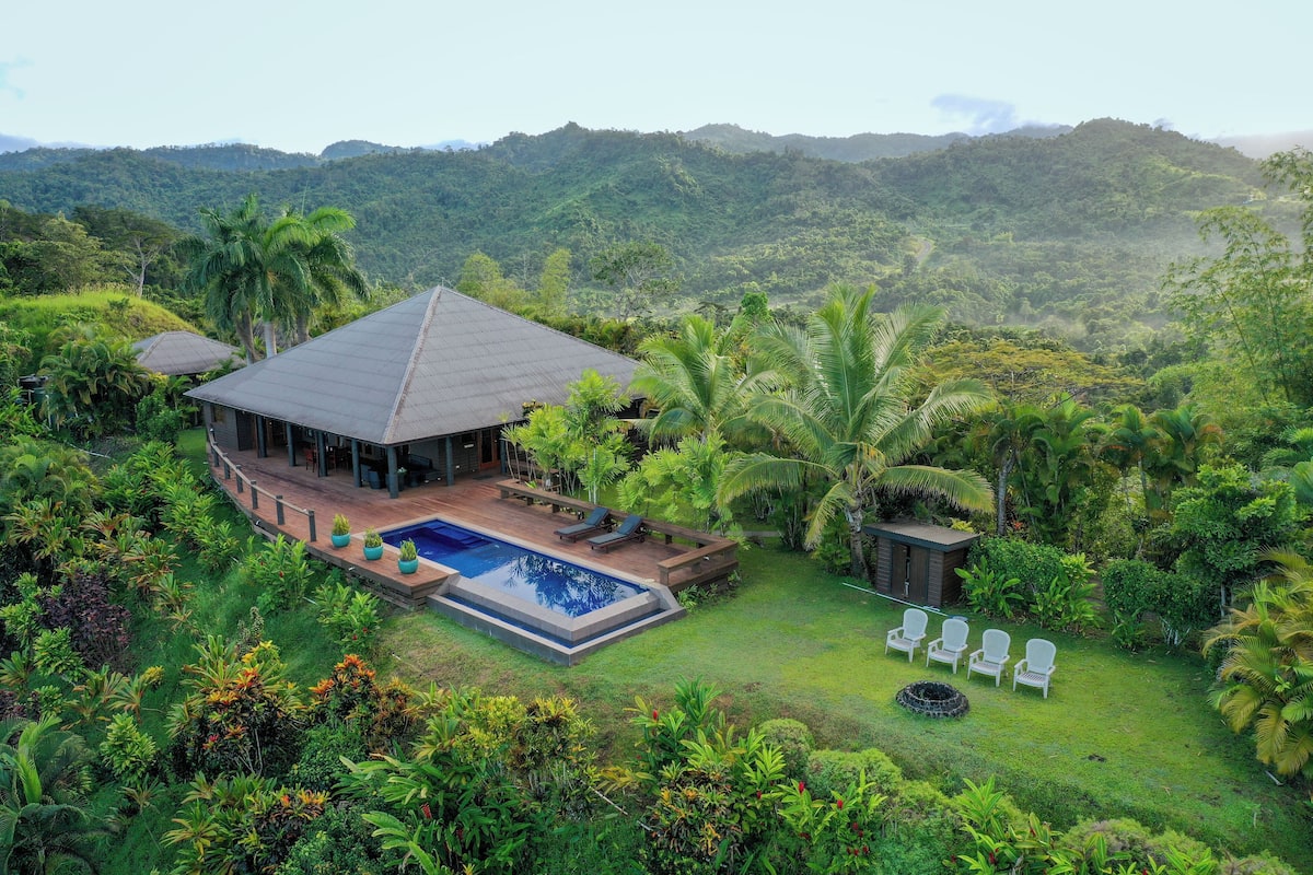 Uma casa com piscina, cercada por vegetação exuberante e colinas ao fundo.