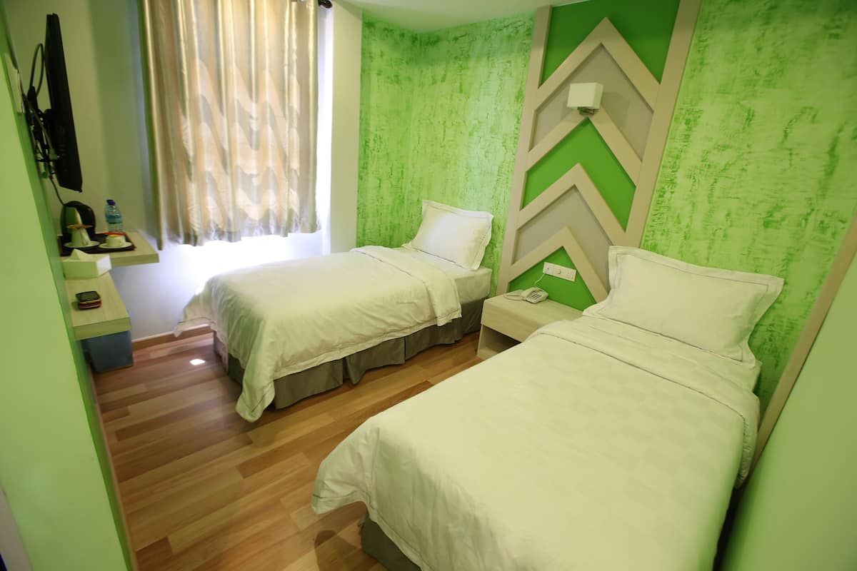 A hotel room with two beds, a wooden floor, a green accent wall, a window with curtains, a bedside table with a phone, and a wall-mounted TV.