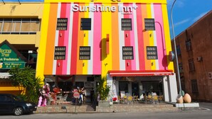 Un bâtiment de l’hôtel Sunshine Inn+, aux couleurs vives, avec une façade à rayures, une entrée bien visible et un espace extérieur aménagé pour s’asseoir.