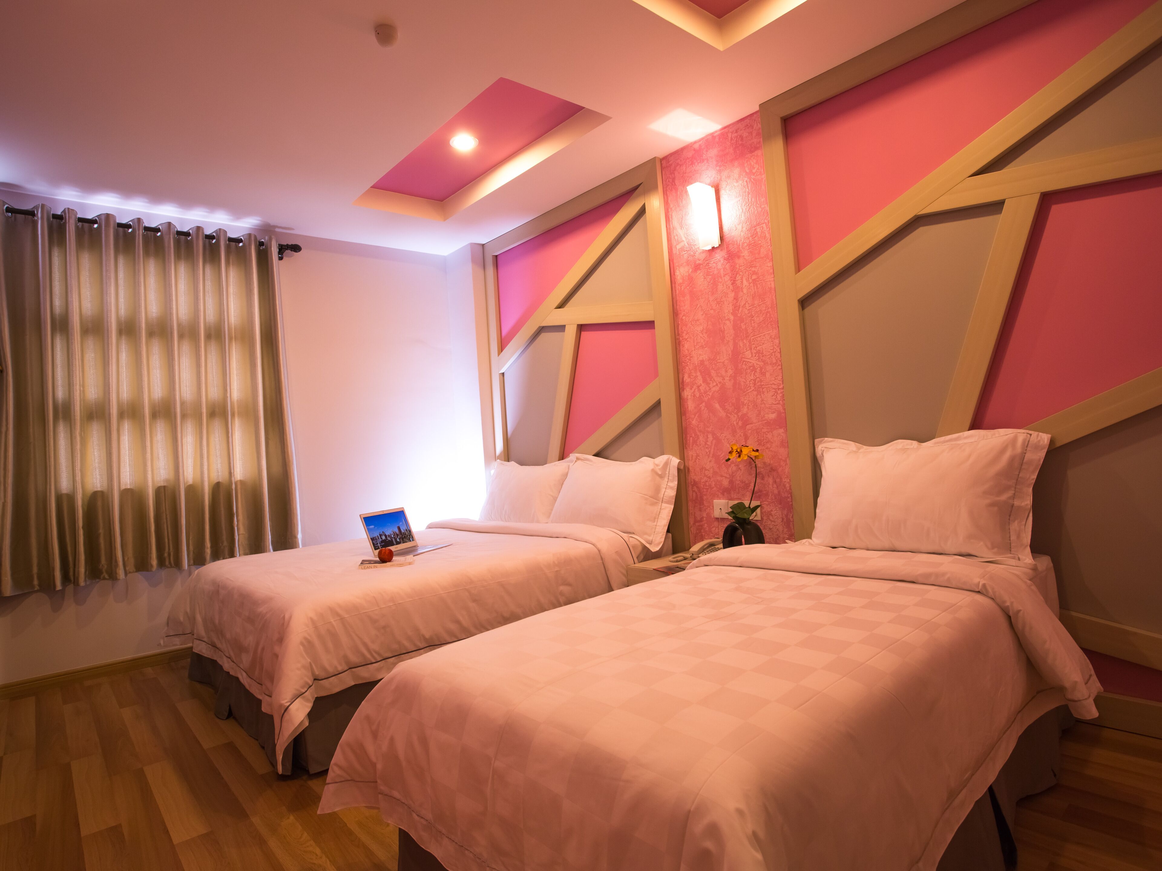 A hotel room with two beds, a window with curtains, a bedside table with a laptop, and a wall with geometric patterns.