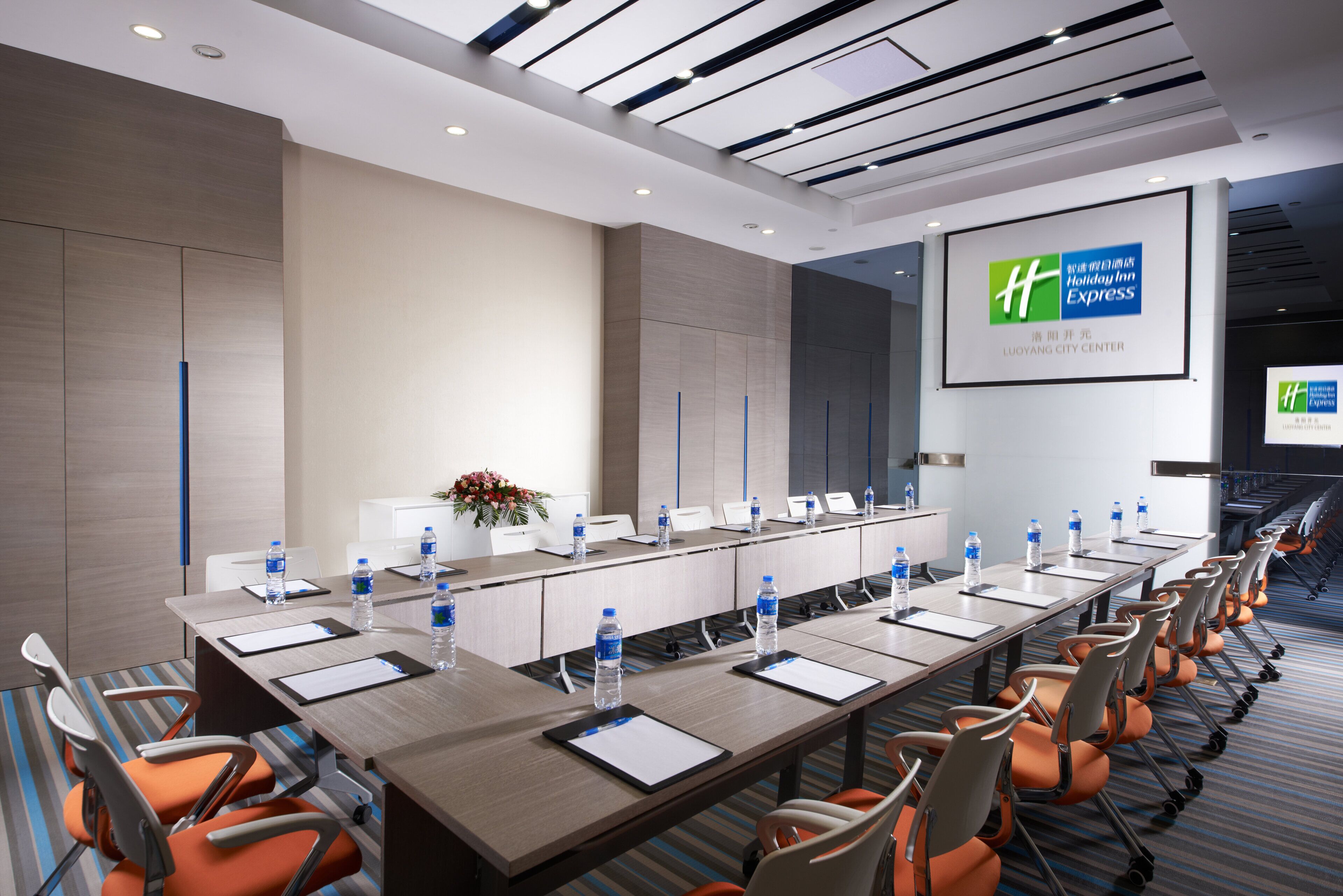 Un salón de conferencias con una mesa larga, sillas, botellas de agua y una pantalla mostrando el logotipo de Holiday Inn Express.
